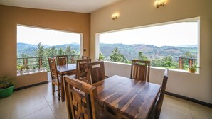 A dining area with a wooden table and chairs, large windows with a view of a mountainous landscape, and potted plants on the balcony. - Penta Peak By Indostays (Kotagiri)