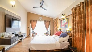 A hotel room with a bed, a desk, a TV, a ceiling fan, and curtains. - Penta Peak By Indostays (Kotagiri)