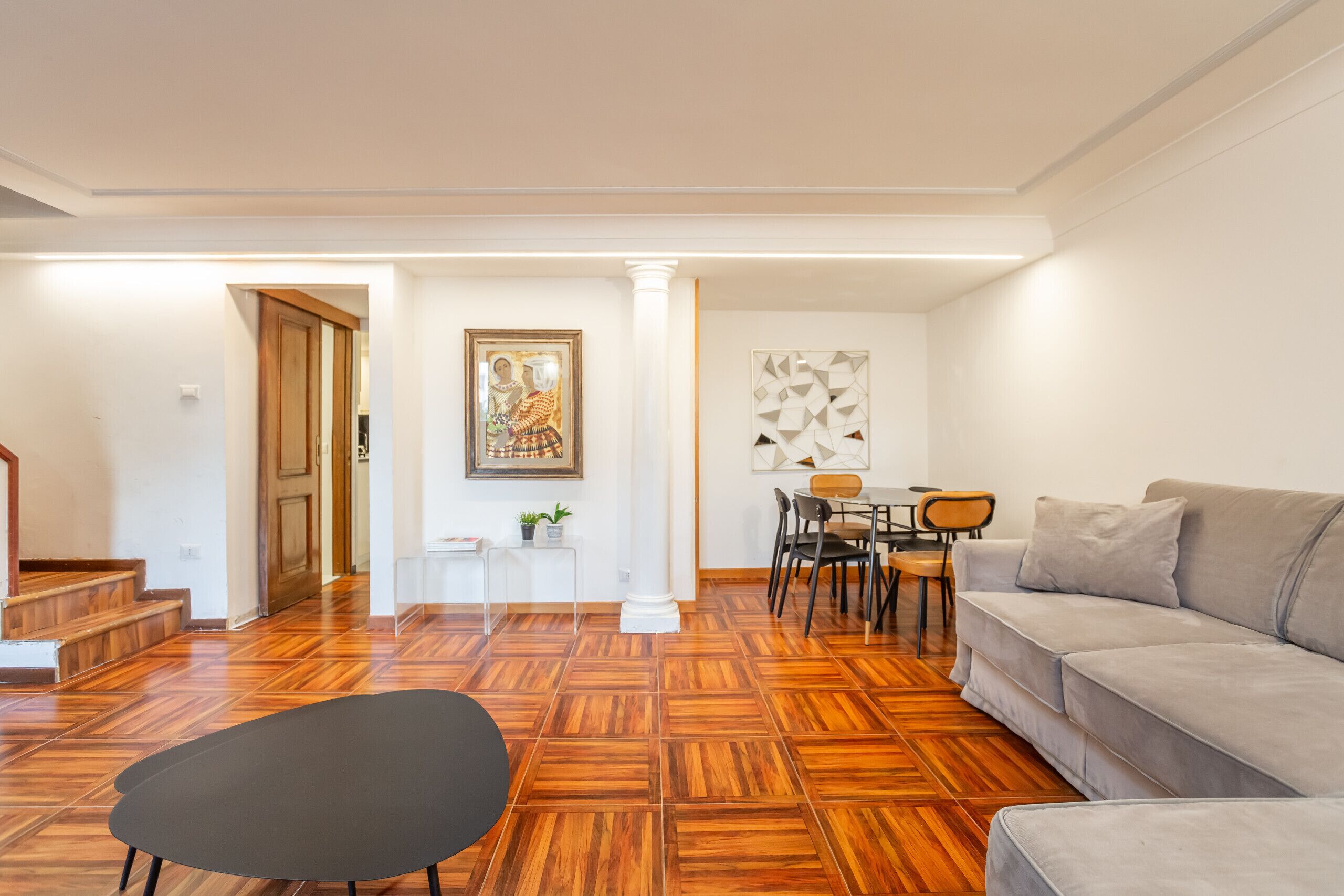Una sala de estar moderna con piso de parquet de madera, un sofá gris, un reposapiés negro, una zona de comedor con mesa y sillas, y una escalera que lleva a otra habitación.