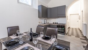 Un coin repas avec une table dressée pour six personnes, une cuisine en arrière-plan et un sol à carreaux.
