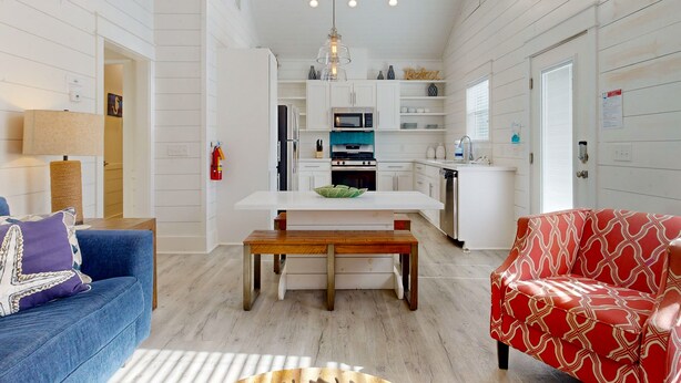 Uma sala espaçosa com um banco de madeira, um sofá azul e um sofá vermelho estampado. Cozinha com armários brancos e uma ilha central visível ao fundo.