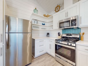 A modern kitchen with stainless steel appliances, white cabinetry, a wooden ceiling, and decorative elements like a ship's wheel and a shelf with glassware.