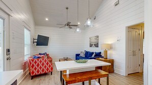 Ein Zimmer mit weißer Holzvertäfelung, einem Deckenventilator, einem flachbildfernseher an der Wand, einem Esstisch mit einer Bank, einem blauen Sofa mit gemusterten Kissen und einem Holznachttisch mit einer Lampe.
