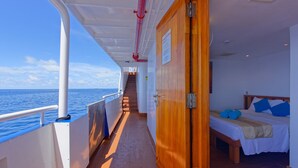 A corridor on a cruise ship with a wooden door leading to a bedroom with a bed, pillows, and a bench. - Yasawa Princess (Malé)