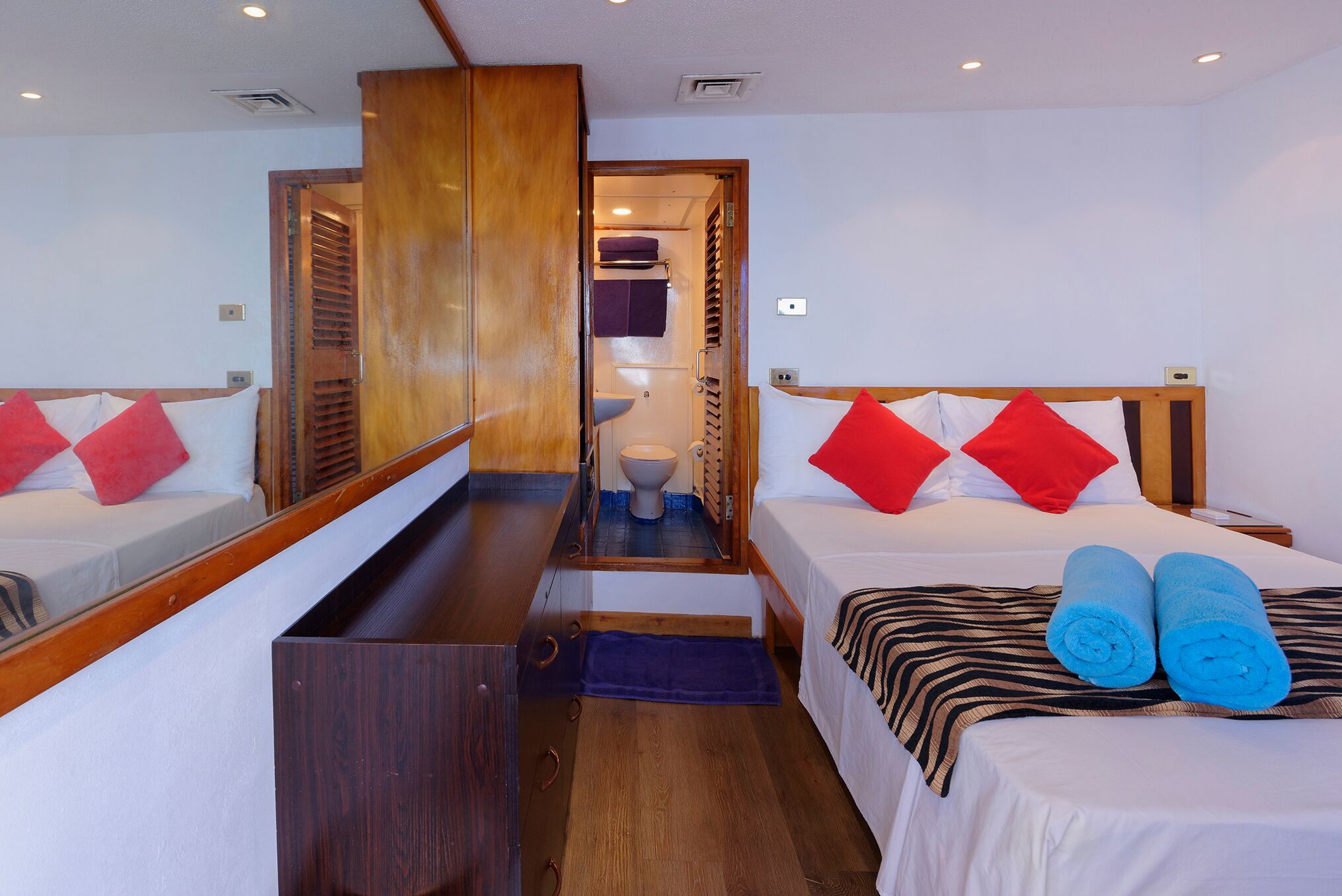 A hotel room with two beds, a wooden wardrobe, a bathroom with a toilet and sink, and a small mirror.