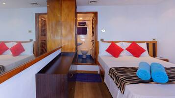 Quarto de hotel com duas camas, um guarda-roupa de madeira, banheiro com vaso sanitário e pia, e um espelho pequeno.
