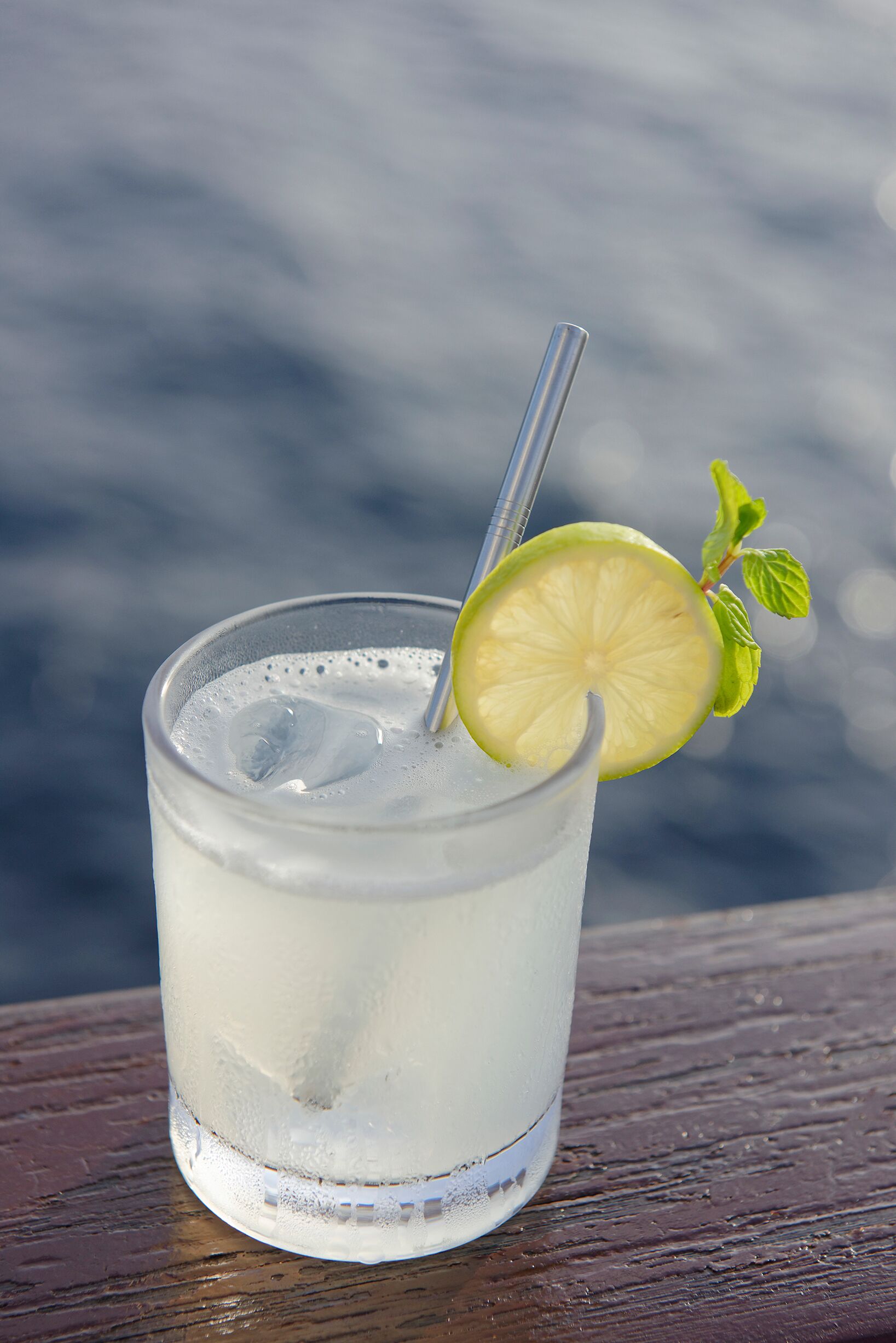 A glass of a clear beverage with a lime slice and mint garnish, placed on a wooden surface with a blurred background.