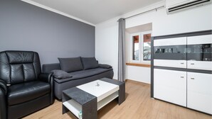 Una sala de estar moderna con un sillón de cuero, sofá gris y una mesa de centro blanca. A la derecha, se observa un aparador con cristalería.