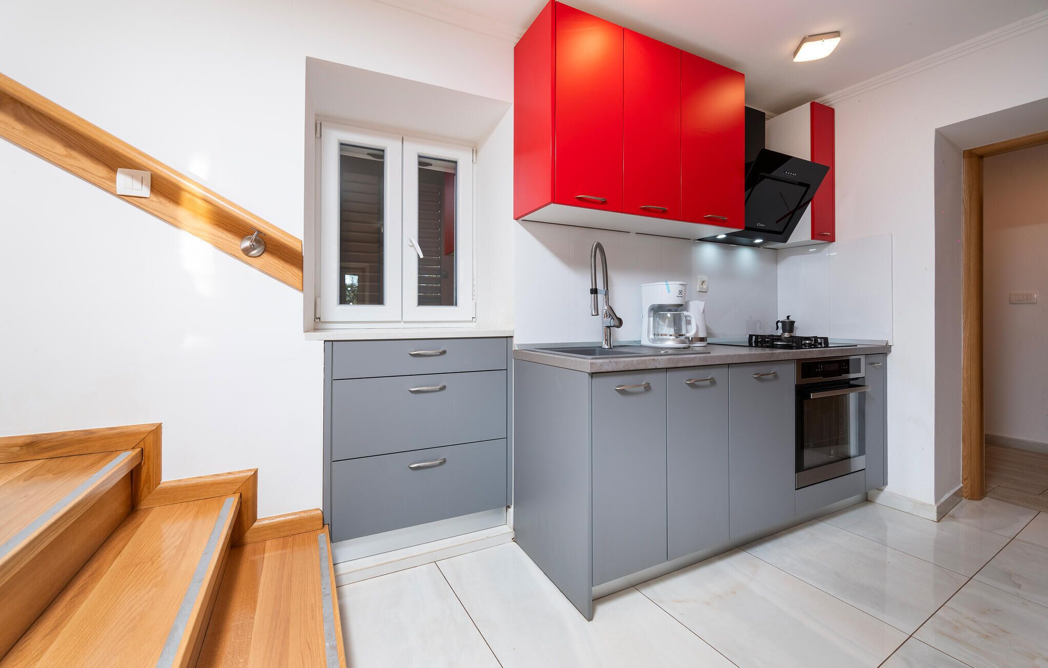 A modern kitchen with grey cabinets, a red upper cabinet, a window, and a built-in oven.