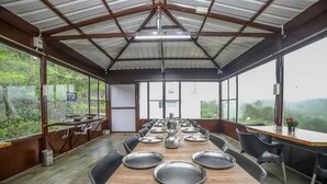A long dining table with metal plates and a pitcher, surrounded by black chairs in a covered dining area with large windows. - Peri Resorts by Indostays (Kotagiri)