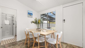 A modern dining area with a wooden table and white chairs, a glass shower enclosure, and a view through a window with blinds. - The Gunyah by Experience Jervis Bay (Huskisson)