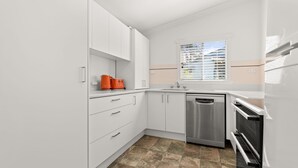A modern kitchen with white cabinets, a stainless steel dishwasher, and a window with blinds. - The Gunyah by Experience Jervis Bay (Huskisson)