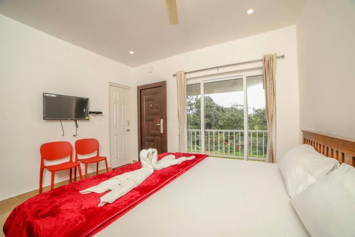 Une chambre d’hôtel avec un lit, une télévision, deux fauteuils rouges, une moquette rouge et un balcon donnant sur un paysage verdoyant.