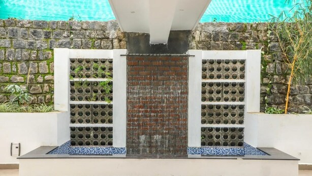 Una fuente exterior moderna con pared de ladrillo y aberturas circulares, rodeada por una estructura blanca y un muro de piedra.