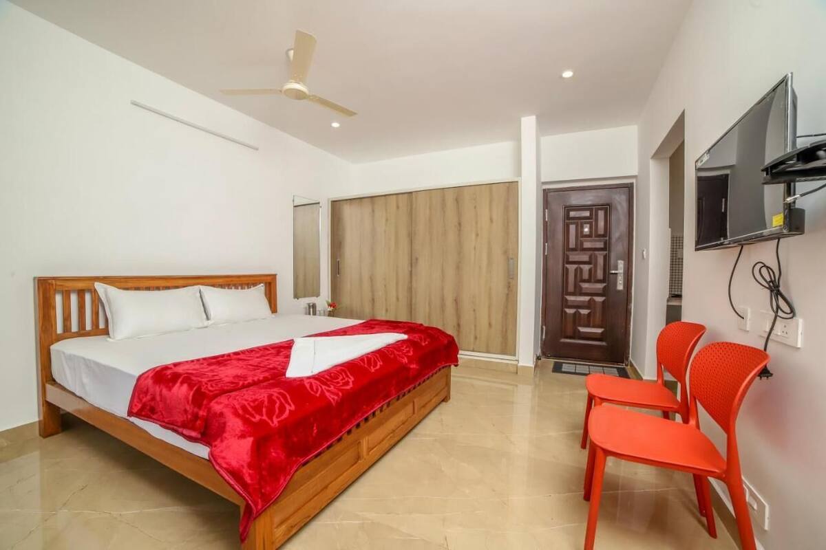 Une chambre avec un lit en bois, une couvre-lit rouge, un ventilateur de plafond, un téléviseur fixé au mur et une porte donnant sur une autre pièce.