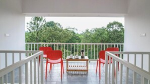 Un balcón con barandilla blanca, dos sillas rojas, una mesita de madera y vistas a árboles verdes.
