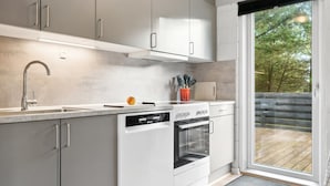 A modern kitchen with grey cabinets, a dishwasher, an oven, and a large window offering a view of a wooden deck.