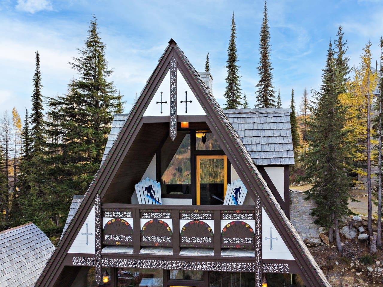 A chalet with a triangular roof, wooden balcony with chairs, and a view of the surrounding forest.