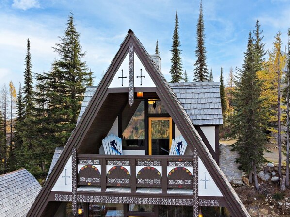 Una cabaña con techo triangular, balcón de madera con sillas y vistas al bosque circundante.
