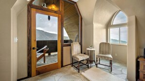 A room with a stone floor, two upholstered chairs, a small table with a plant, and a wooden door leading to an outdoor area with a view of mountains. - Ski-In/Ski-Out Schweitzer Chalet + Private Hot Tub - Guest Favorite (Sandpoint)