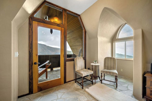 Une pièce dotée d’un sol en pierre, de deux fauteuils rembourrés, d’une petite table ornée d’une plante et d’une porte en bois donnant sur un espace extérieur avec vue sur les montagnes.