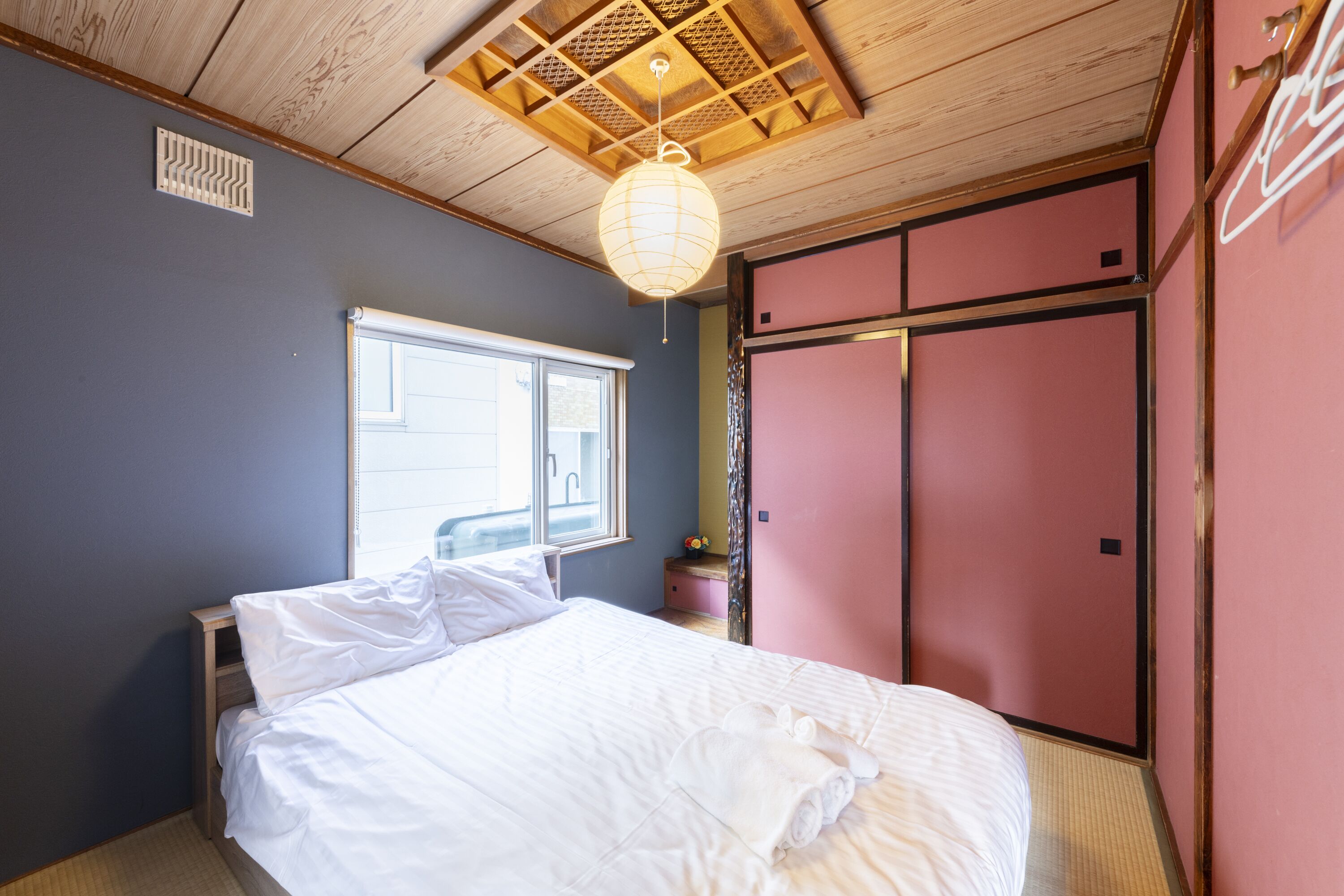 A bedroom with a bed, a window, a wooden ceiling, and a pink wardrobe.
