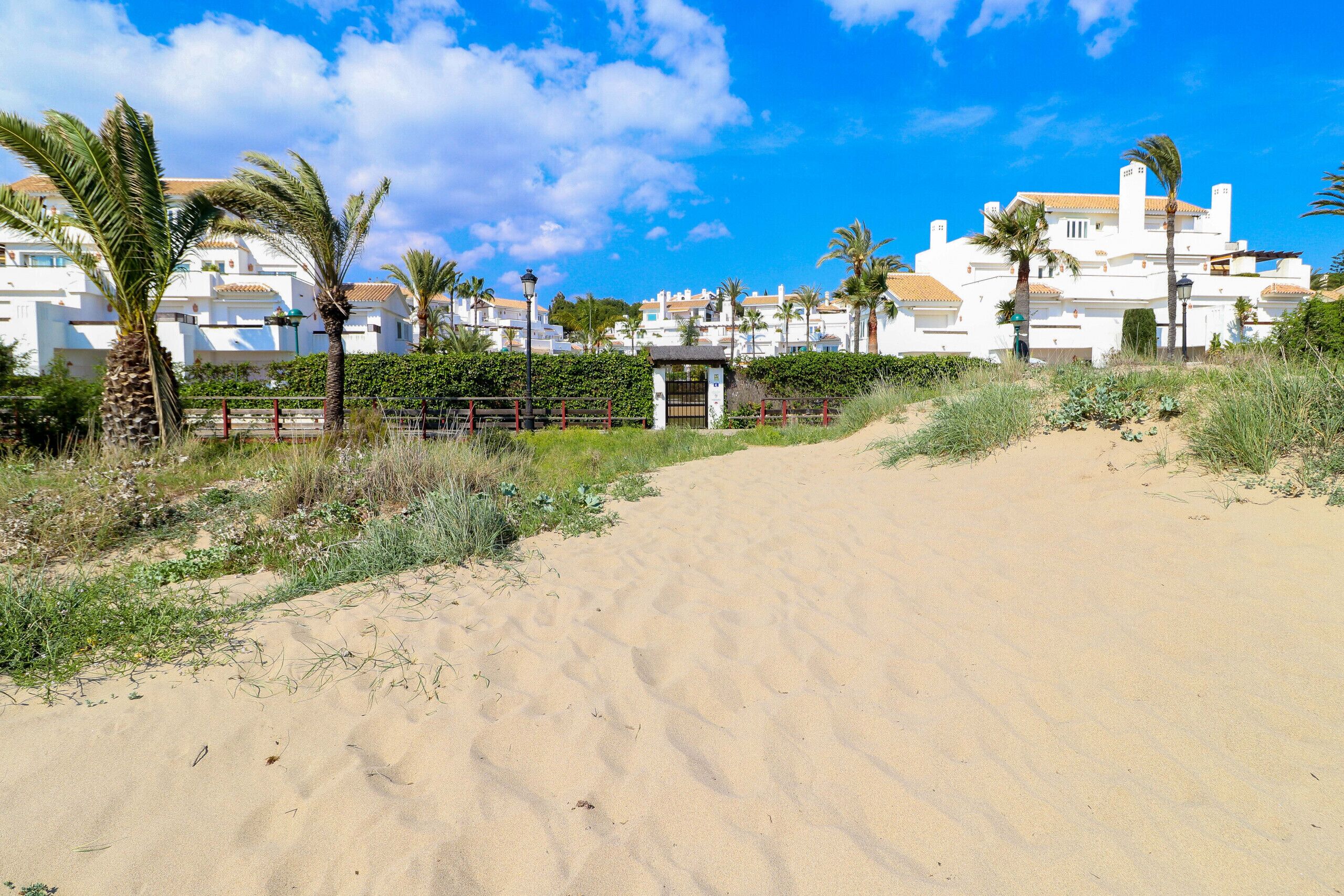 A sandy path leading to a gated entrance with a white building in the background.