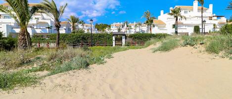 A sandy path leading to a gated entrance with a white building in the background.