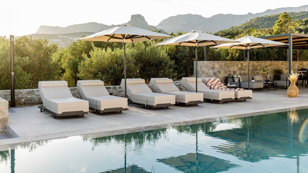 A poolside area with lounge chairs and umbrellas, a stone wall, and a view of mountains. - Beach Villas in Crete Alope & Ava (Ierapetra)