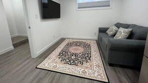 A modern living room with a grey sofa, a flat-screen TV mounted on the wall, and a patterned rug on the floor. - Serene Haven in Harbour Landing (Regina)