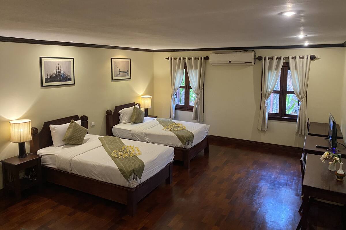 Une chambre d’hôtel avec deux lits, un bureau et une chaise. Elle comprend deux fenêtres avec des rideaux, un climatiseur fixé au mur et des tableaux encadrés accrochés au mur.
