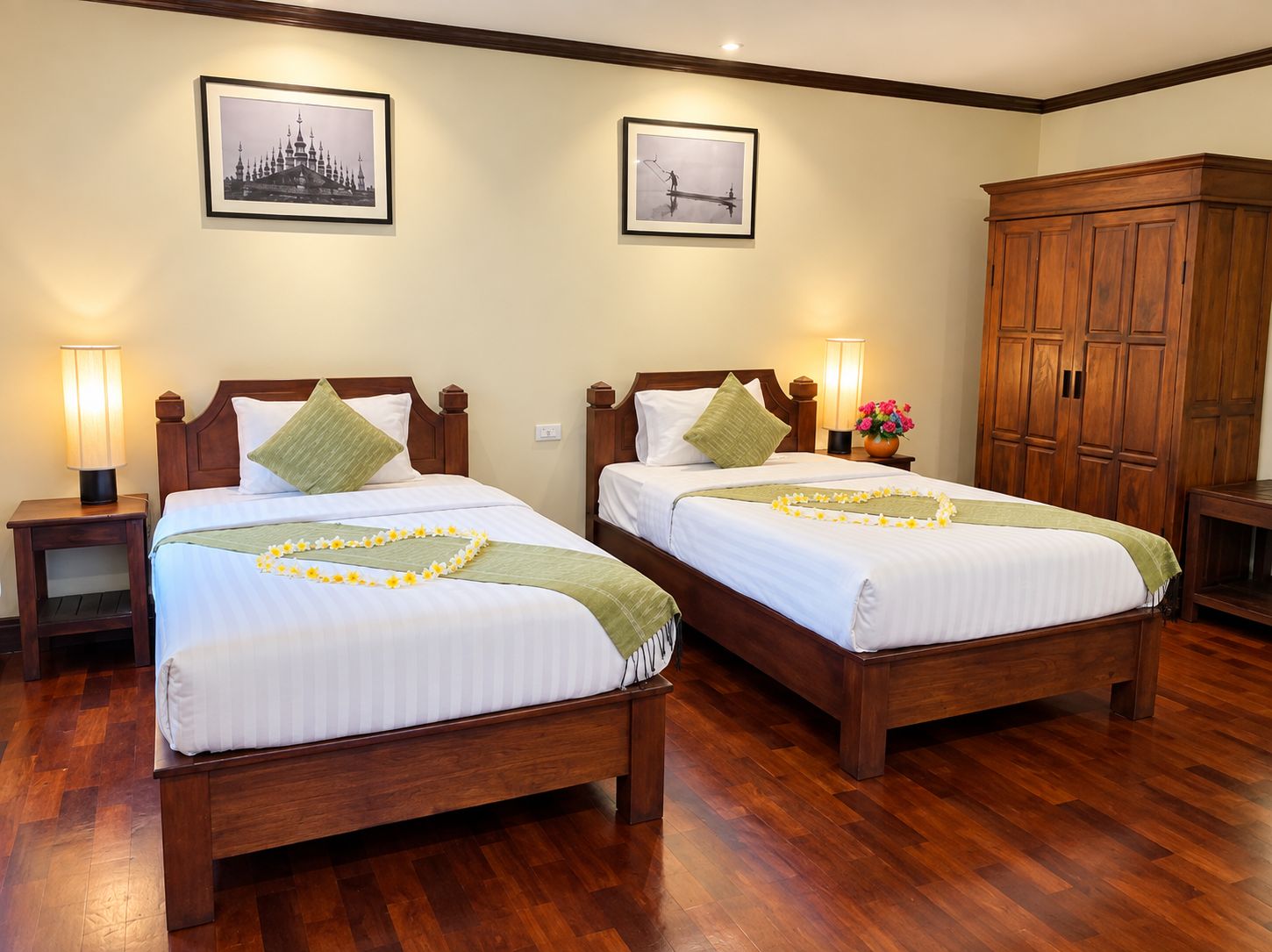 Two wooden beds with white linens, green throw pillows, and floral arrangements. There are framed pictures on the wall and a wooden wardrobe.