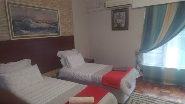 A hotel room with two beds, a painting on the wall, an air conditioning unit, and a window with curtains. - Comrades Oasis Guest House  (Pietermaritzburg)