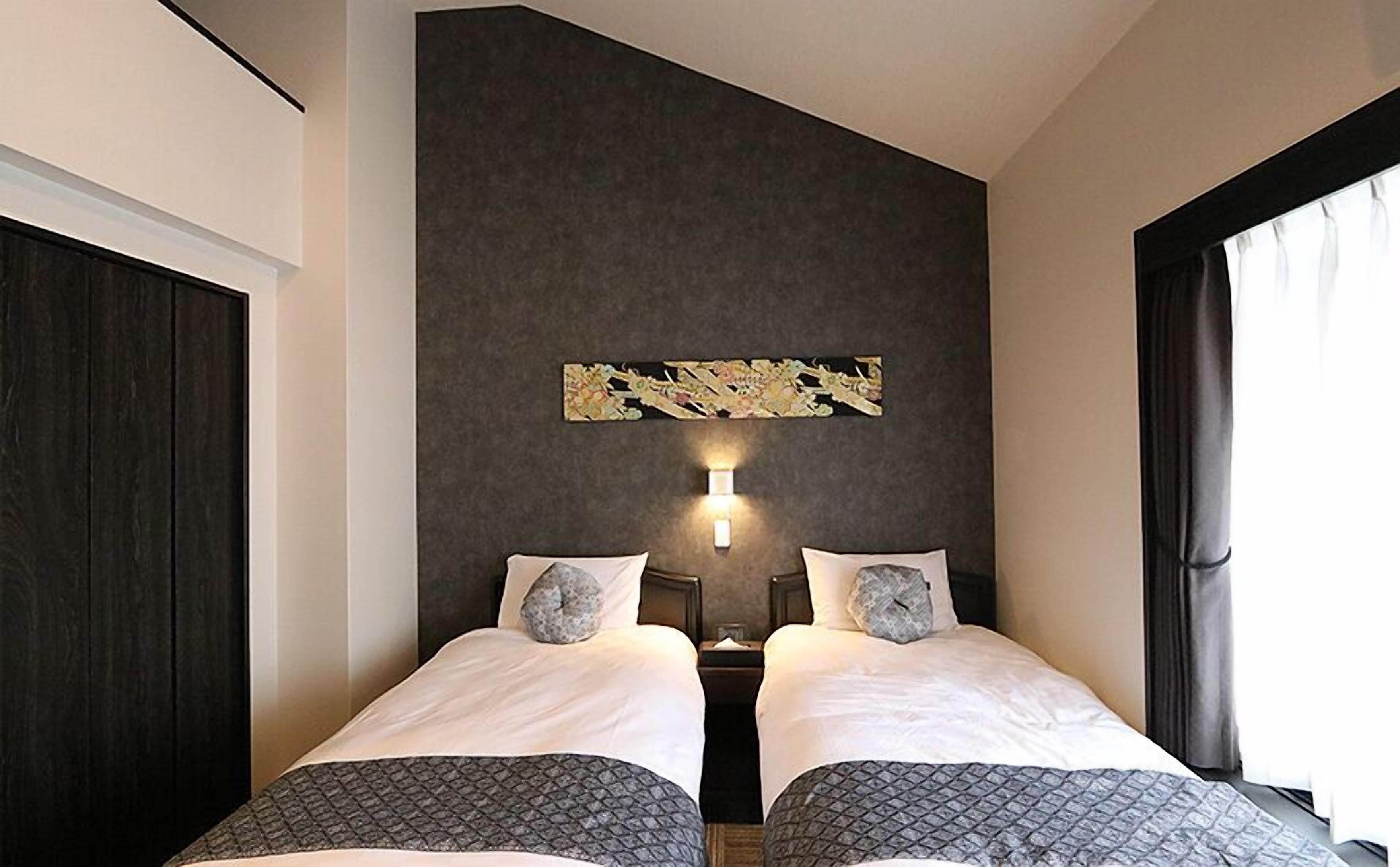 A hotel room with two beds, a dark wall, a decorative wall piece, and a nightstand with a lamp.