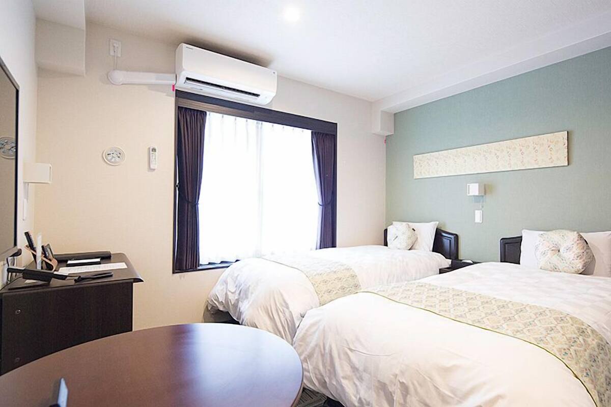 Une chambre d’hôtel avec deux lits, un bureau, un appareil de climatisation, une fenêtre avec des rideaux et un luminaire fixé au mur.