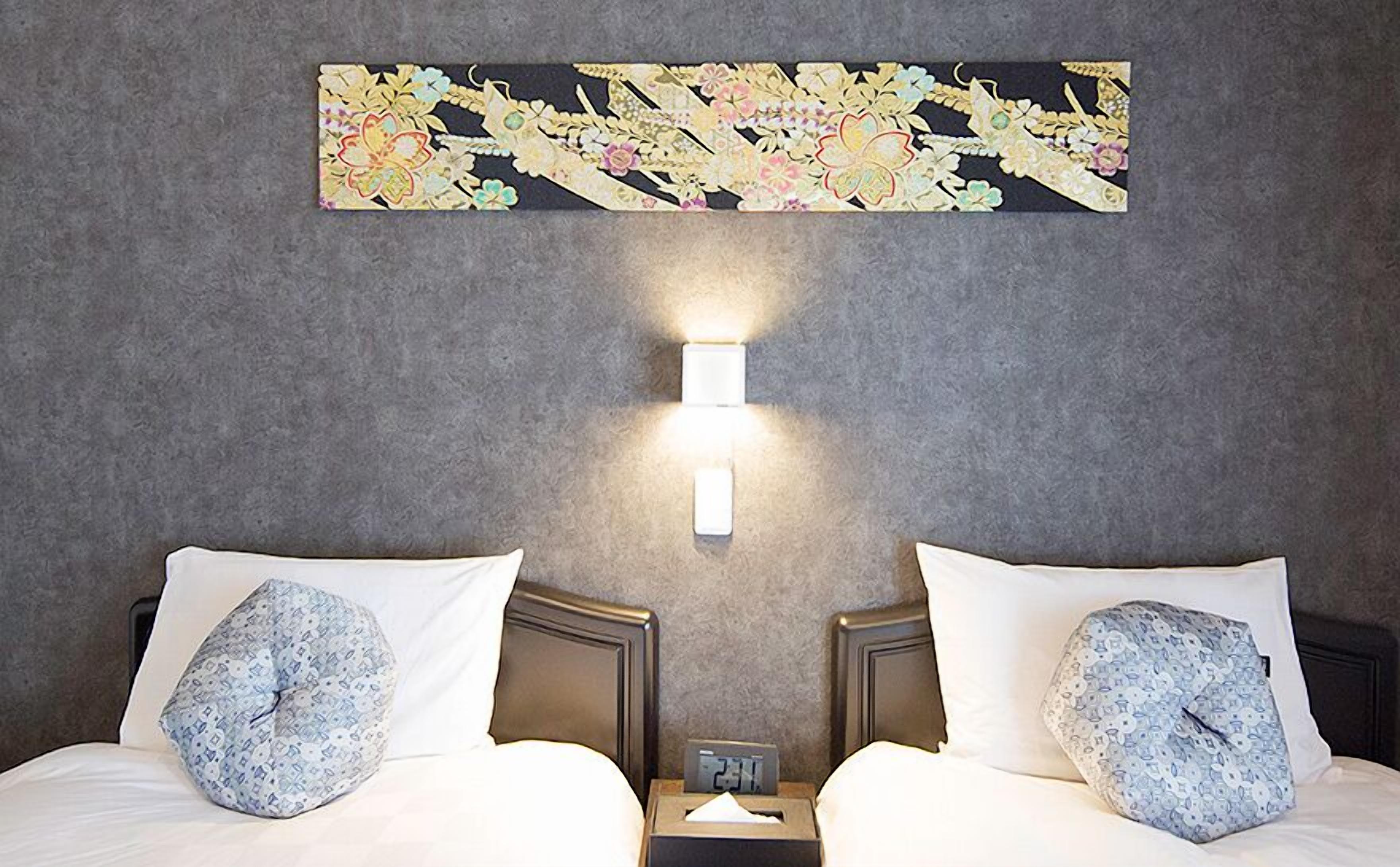 A hotel room with two beds, each with a patterned pillow, a nightstand with a clock, and a wall-mounted light fixture.
