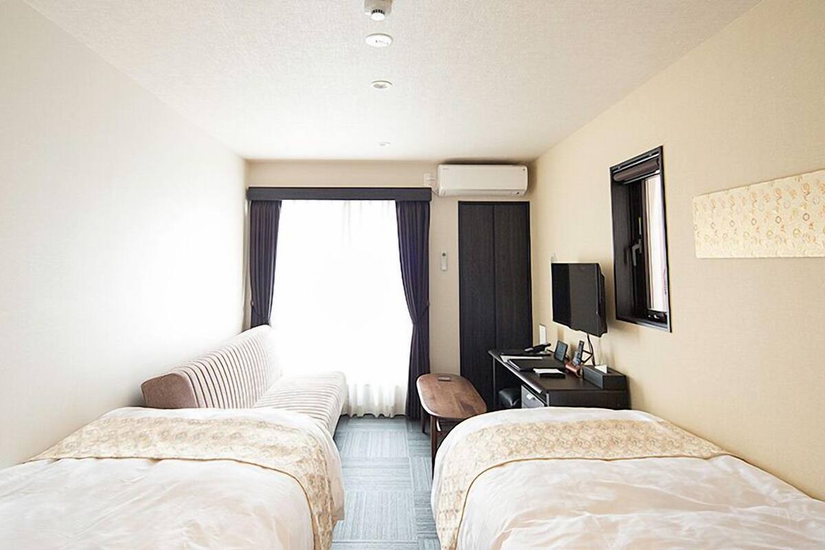 Une chambre d’hôtel avec deux lits, un bureau avec une chaise, un téléviseur à écran plat et une fenêtre avec des rideaux.