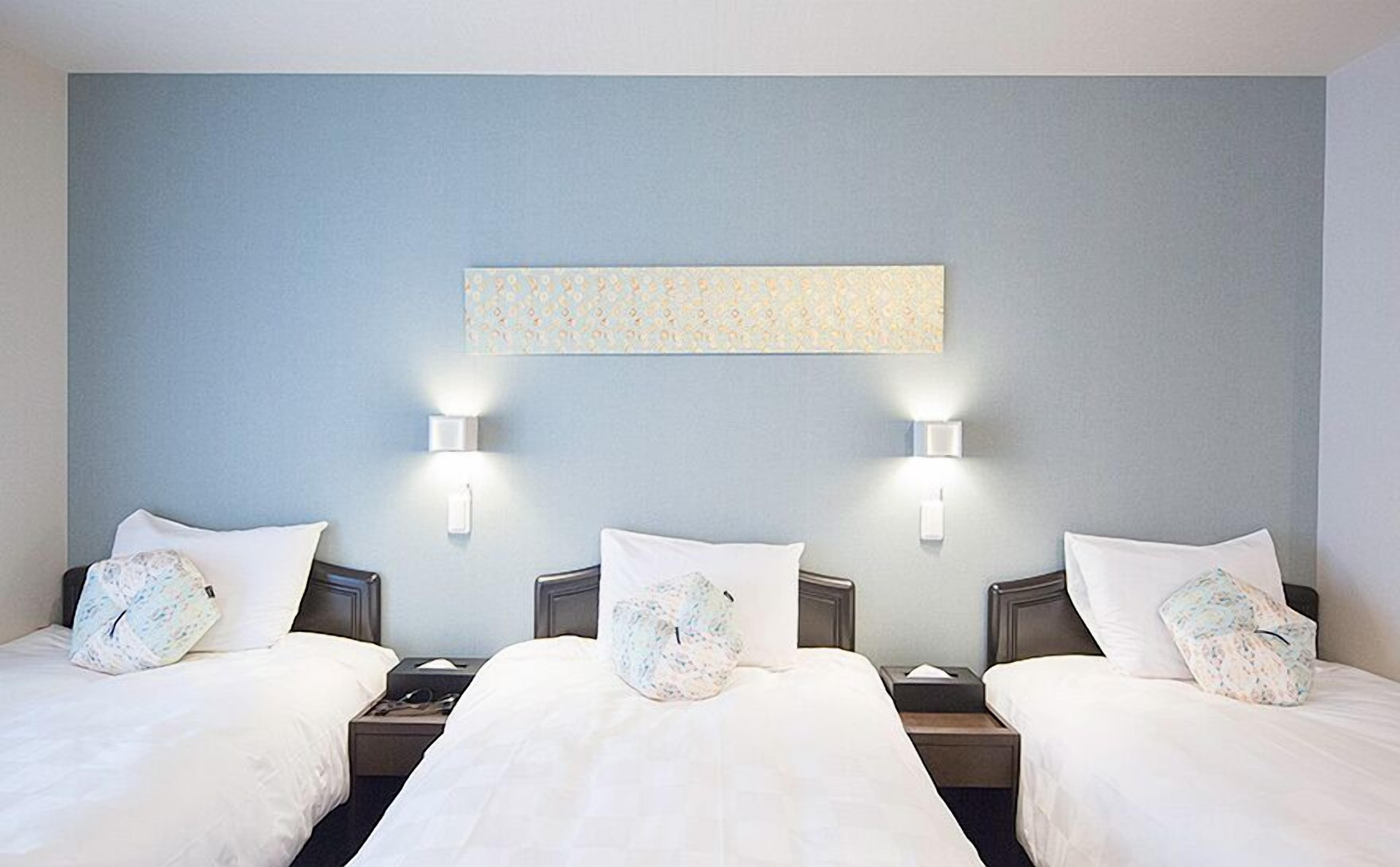 Una camera d'albergo con tre letti, ognuno con biancheria da letto bianca e cuscini coordinati. Due lampade a parete fiancheggiano un pannello decorativo.