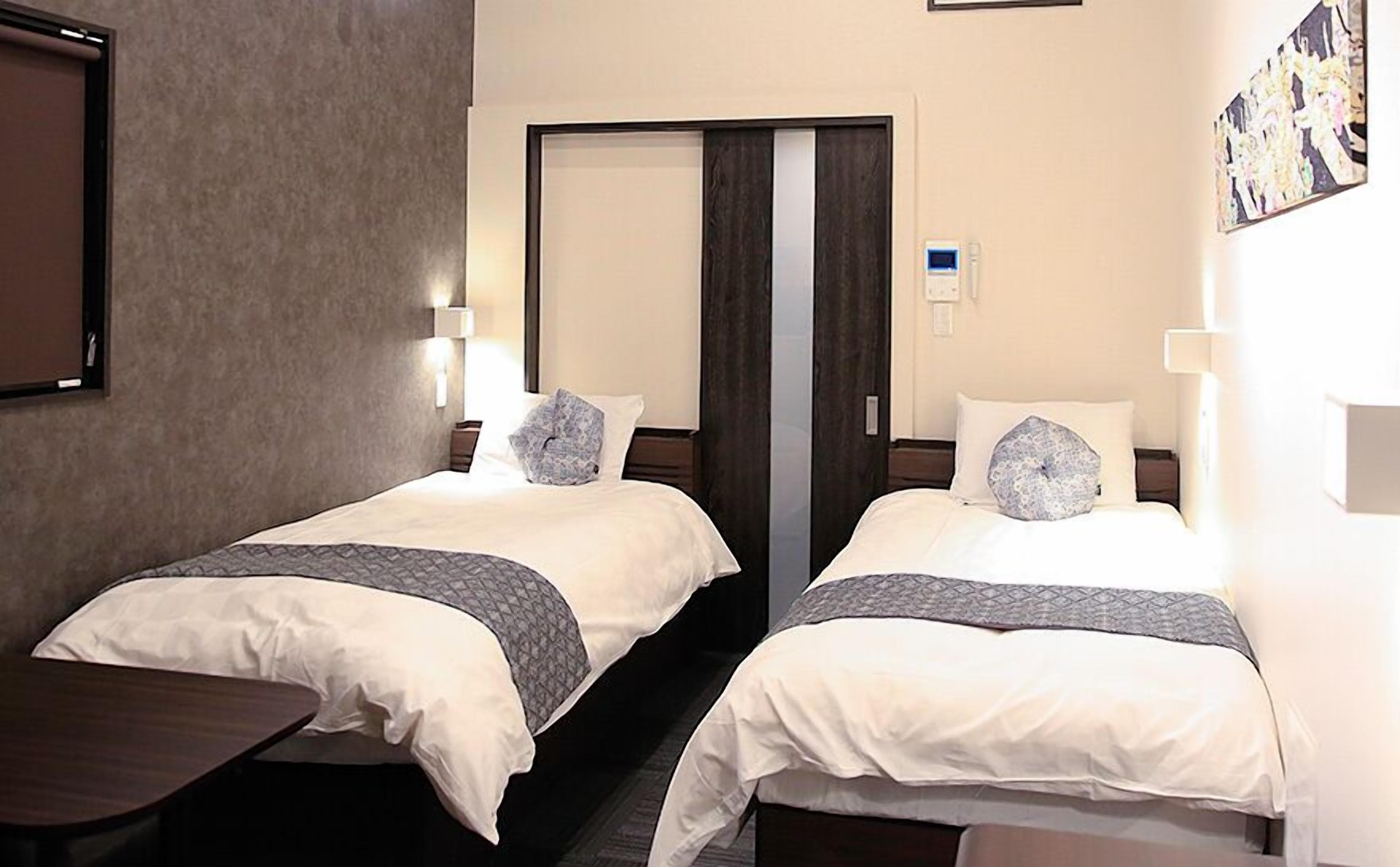 A hotel room with two beds, each with a pillow and a blanket. There is a bedside table and a mirror on the wall.