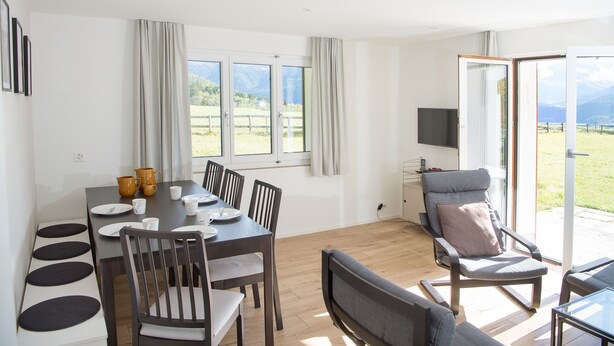 Un coin repas avec une table dressée pour quatre personnes, un téléviseur fixé au mur et une vue sur un champ verdoyant par les fenêtres.