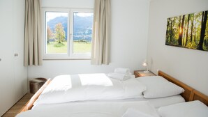 Une chambre avec deux lits, une fenêtre avec des rideaux, un tableau représentant une forêt et une table de chevet avec une lampe.