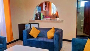 A living room with a blue sofa and armchairs, a coffee table, and a mirror above a kitchen area.