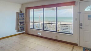 Habitación con un amplio ventanal que da a una playa, una puerta con un letrero y una estantería con diversos objetos.