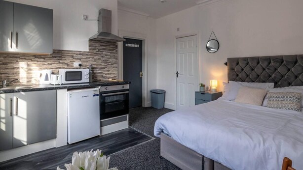 A modern kitchen with a microwave, toaster, and oven, a bedroom with a large bed and nightstand, and a door leading to another room. - Lovely 2-bed Studio10 in Coventry (Coventry)