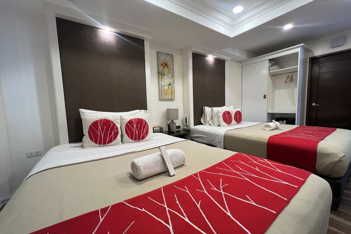 A hotel room with two beds, each with a red and white patterned bedspread, a white towel, and matching pillows. There is a painting on the wall, a bedside table with a lamp, and a wardrobe with a mirror.