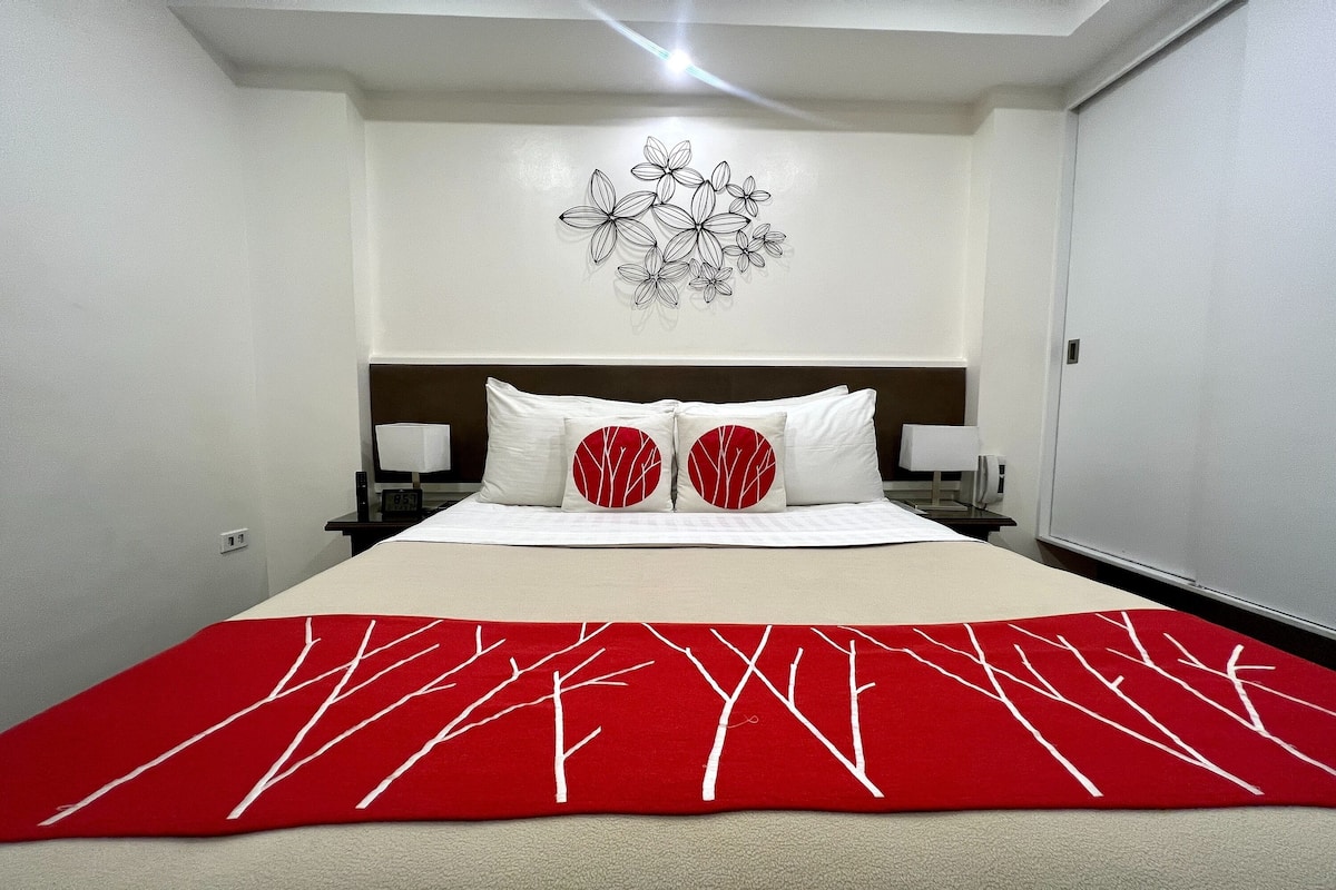 A neatly made bed with a red and white patterned bedspread, a wall art of flowers, and a nightstand with a lamp.