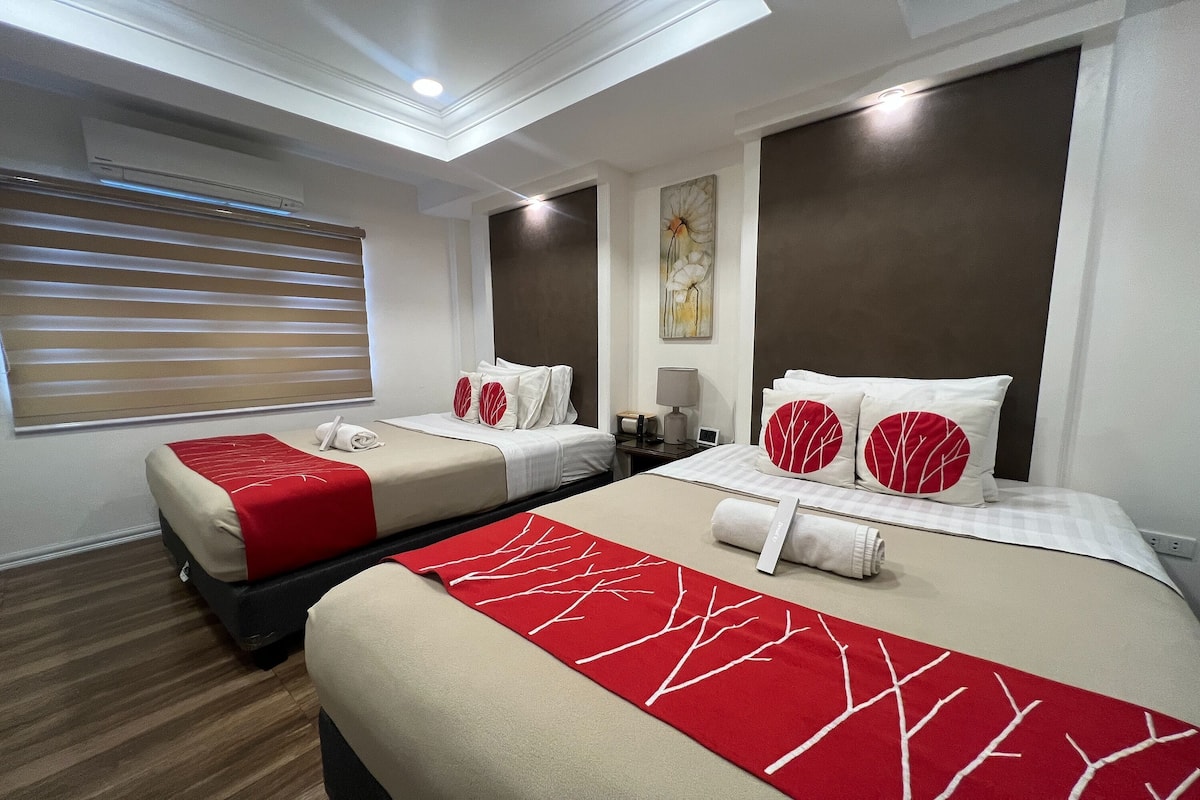 A hotel room with two beds, each with red and white bedding, a large window with blinds, a ceiling light, and a wall-mounted air conditioner.