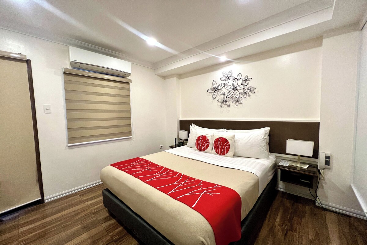 A hotel room with a bed, a nightstand, a wall-mounted air conditioner, a window with blinds, and a decorative wall piece.