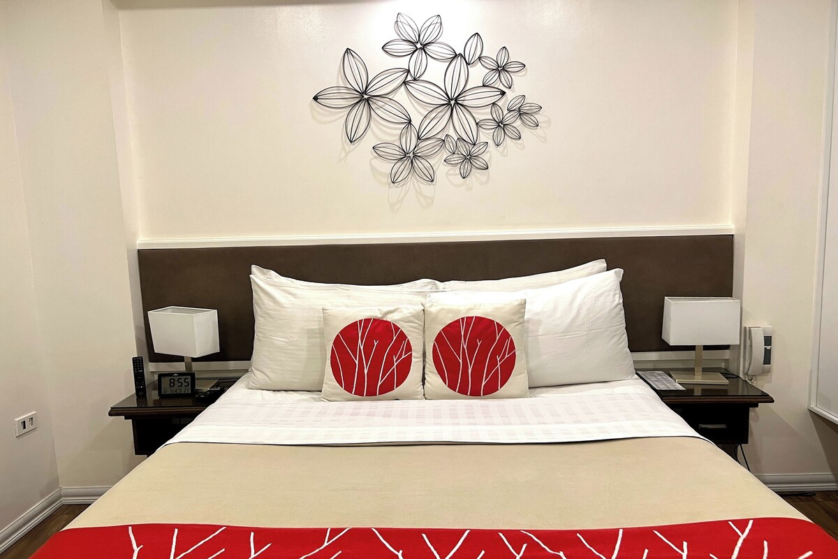 A hotel room with a large bed, two nightstands, a wall art piece, and a red and white patterned throw blanket.