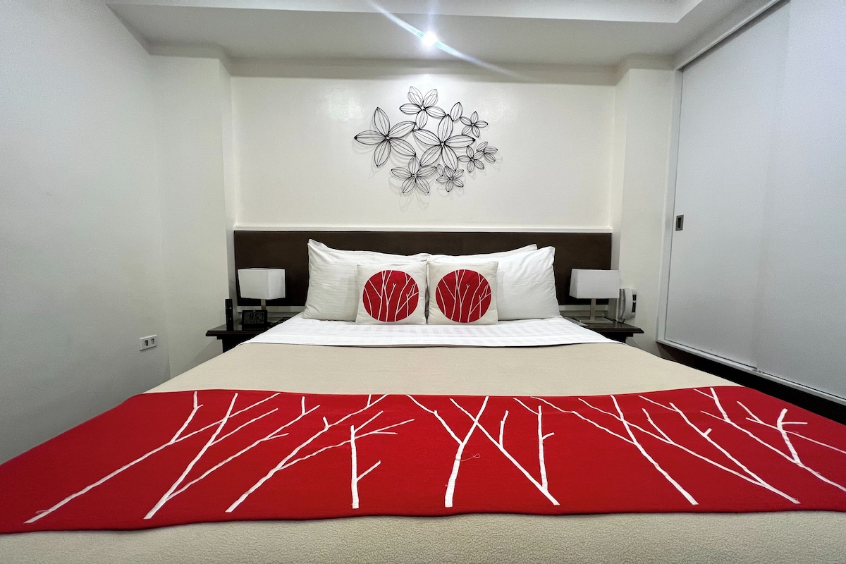 A neatly made bed with a red and white tree-patterned blanket, a wall art of flowers, and two bedside lamps.